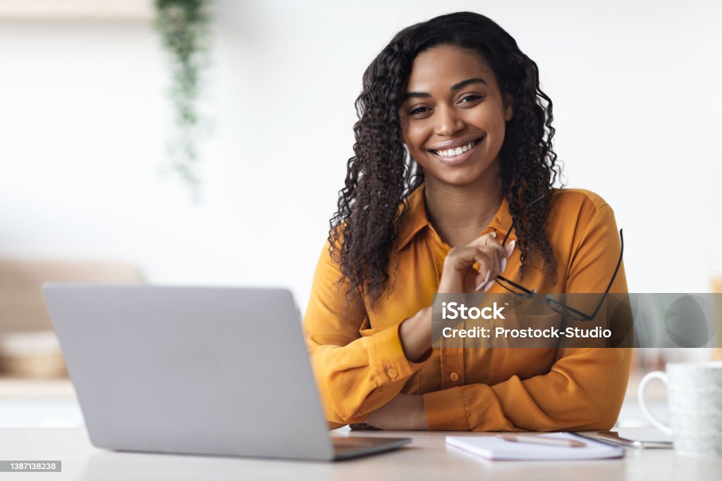 Pretty long-haired millennial african american woman freelancer using laptop at home, black lady sitting at table at kitchen, holding eyeglasses and smiling at camera, copy space. Remote job concept